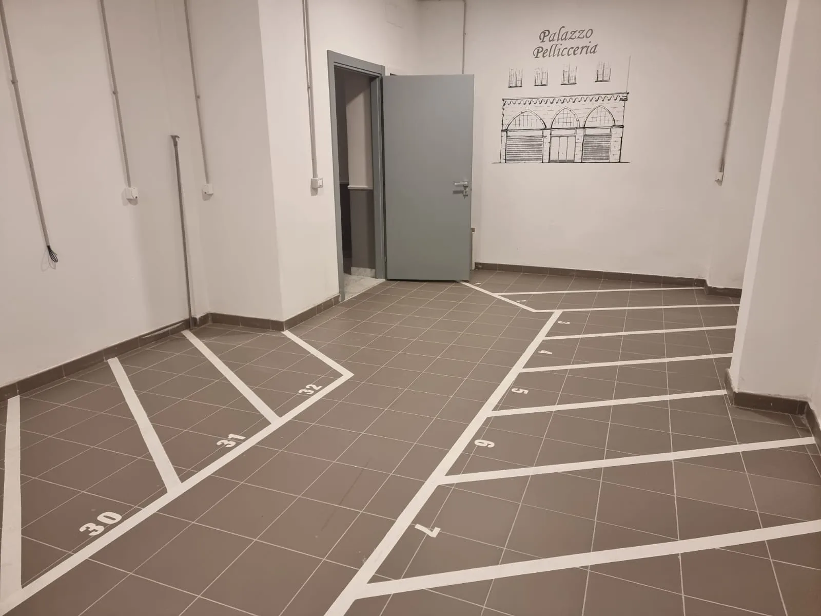 Un espacio interior embaldosado tiene líneas blancas que delimitan varias zonas junto a una puerta abierta.