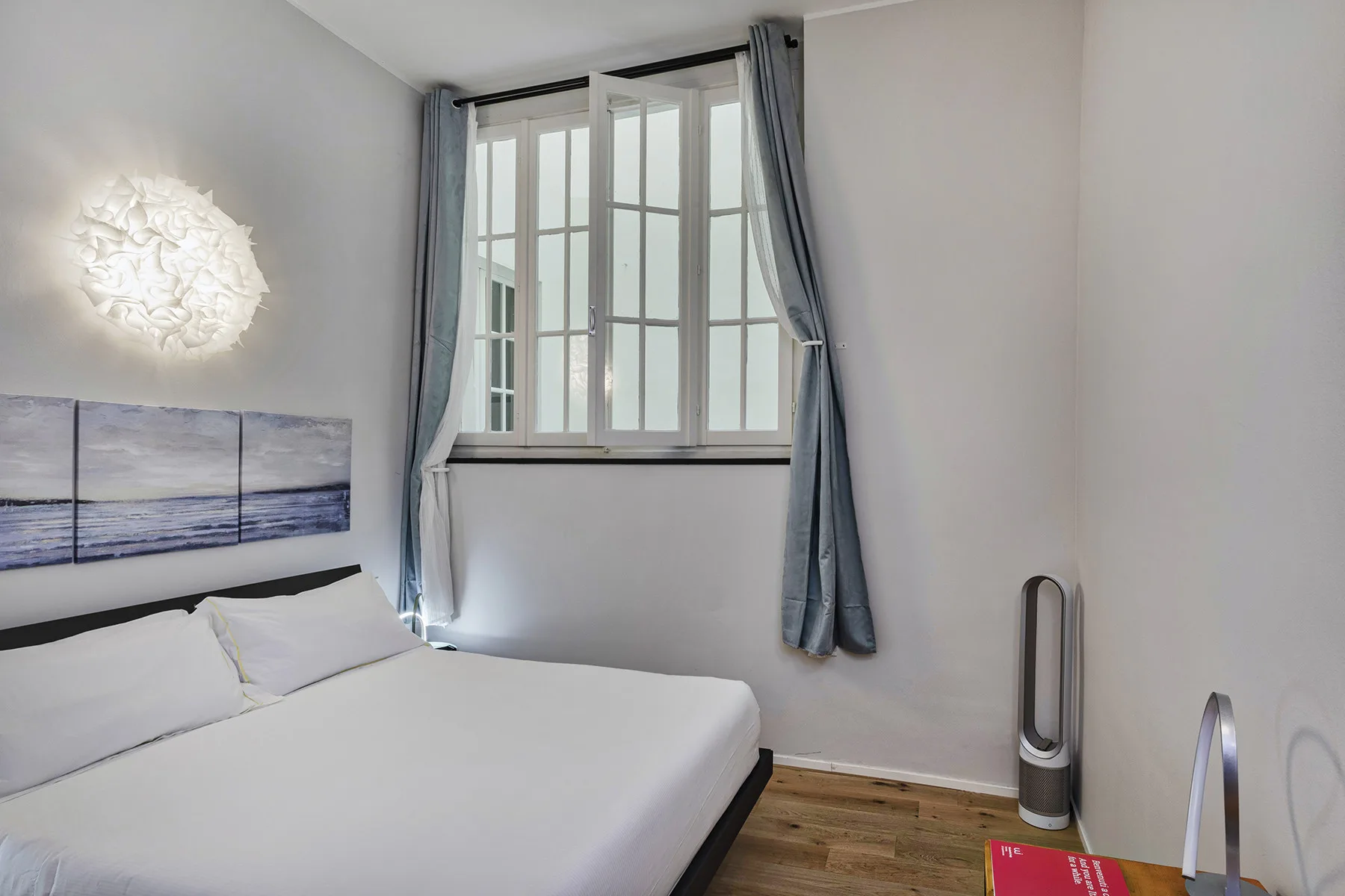 Un dormitorio luminoso con una cama bajo una ventana alta y cortinas grises.