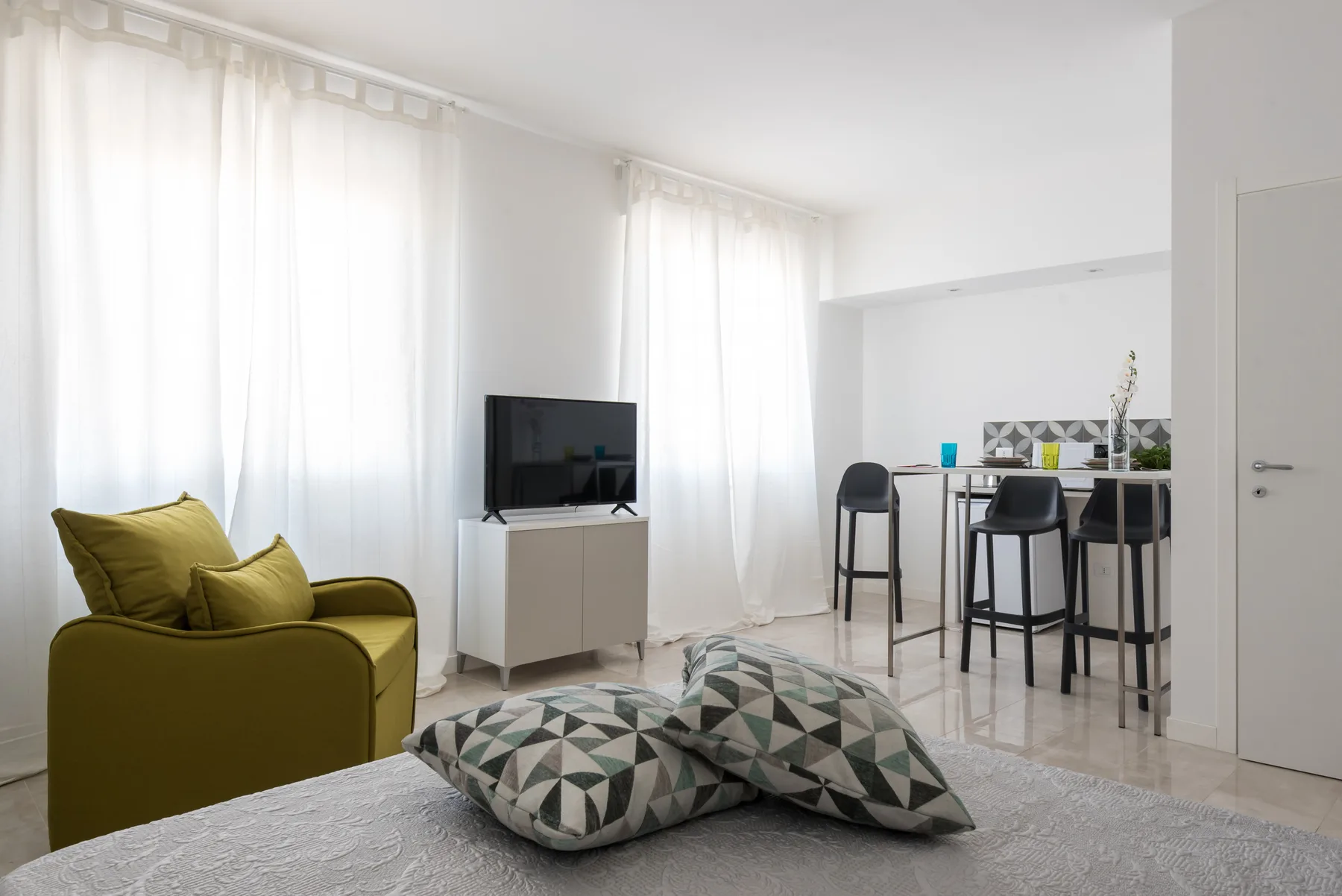 Una sala luminosa con un sillón verde oliva, un televisor y asientos de barra.