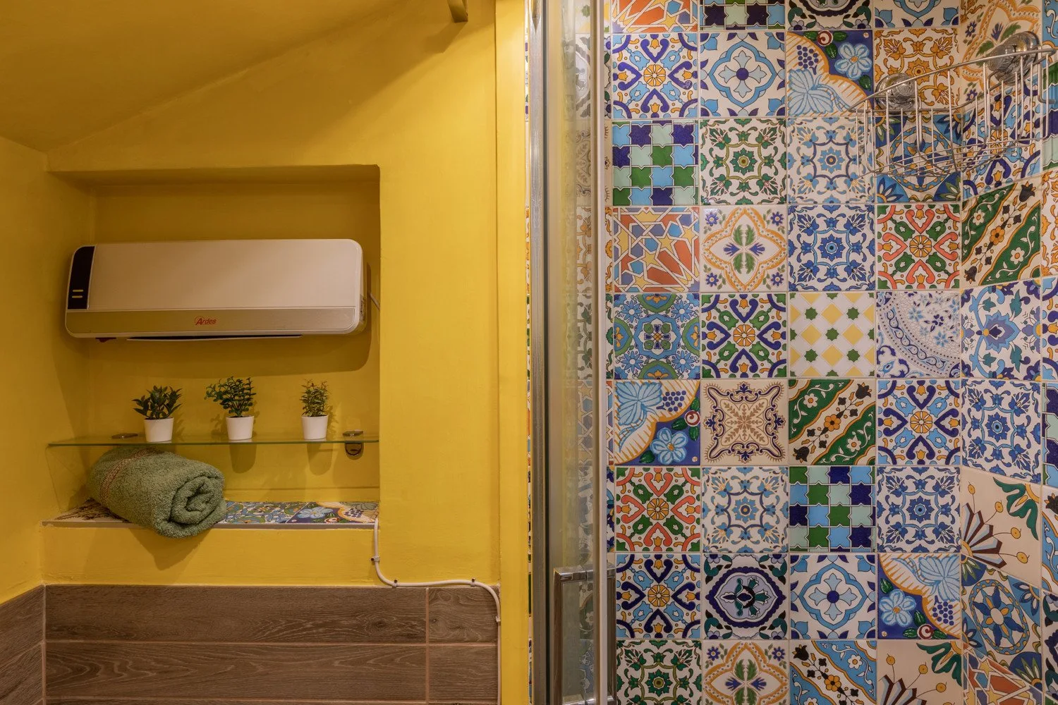 A colorful tiled shower area beside a yellow wall with a towel shelf.