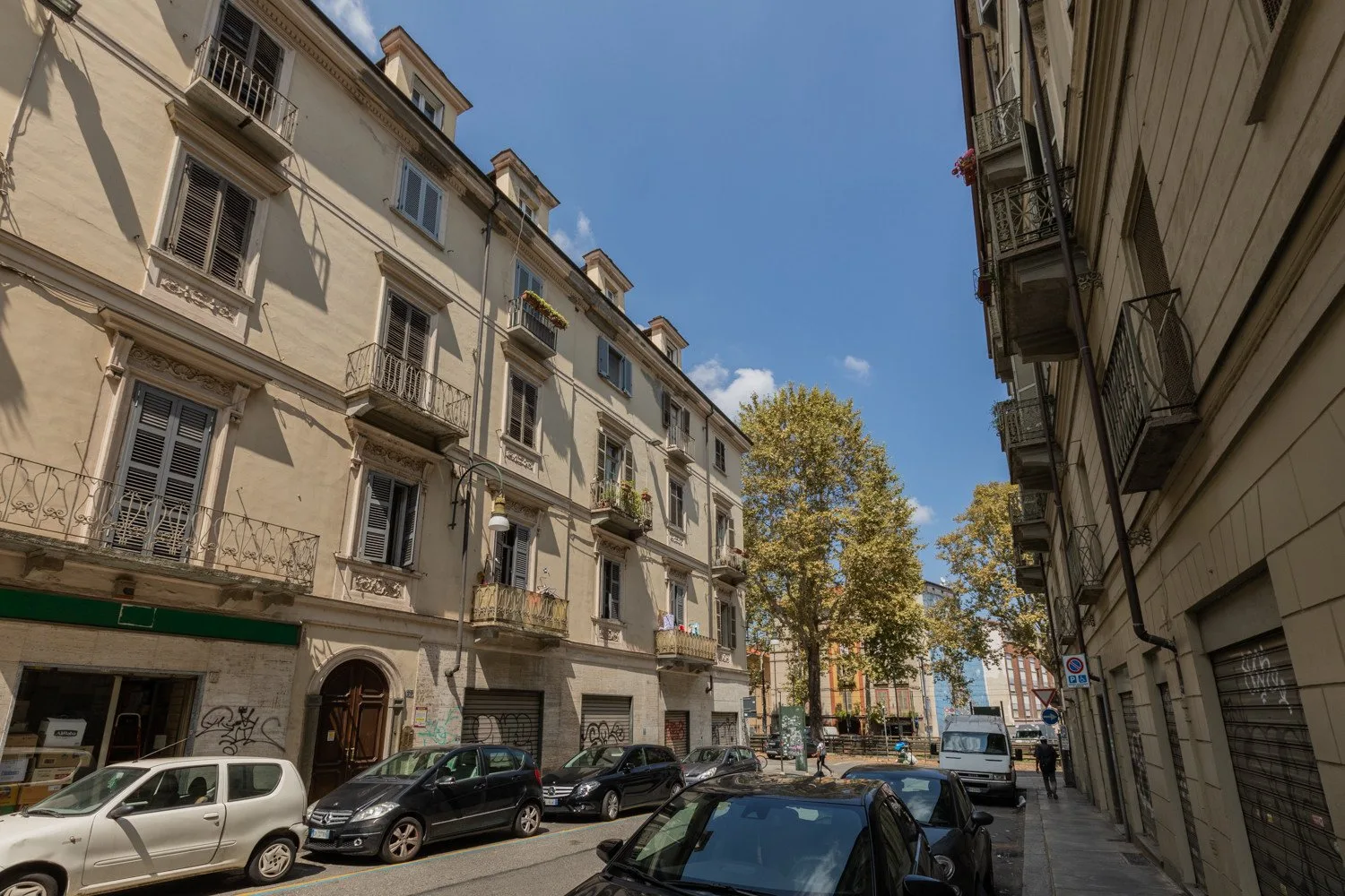 A city street lined with multi-story buildings, parked cars, and trees.