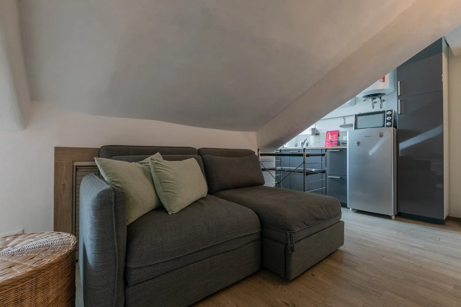 A living area with a sectional sofa beneath a sloped ceiling, opening onto a kitchen.