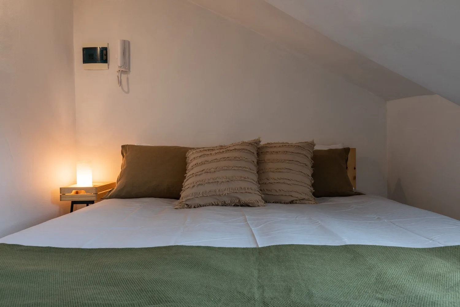 Una cama con varias almohadas está bajo un techo inclinado, junto a una mesita iluminada.