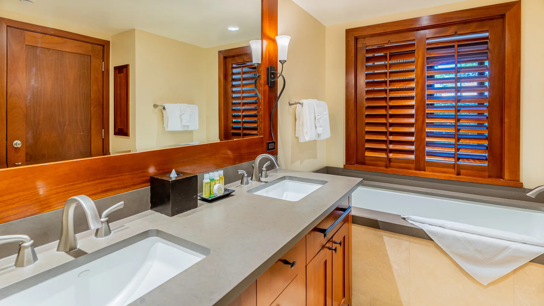 Un tocador de baño con dos lavabos está bajo persianas de madera y un espejo.