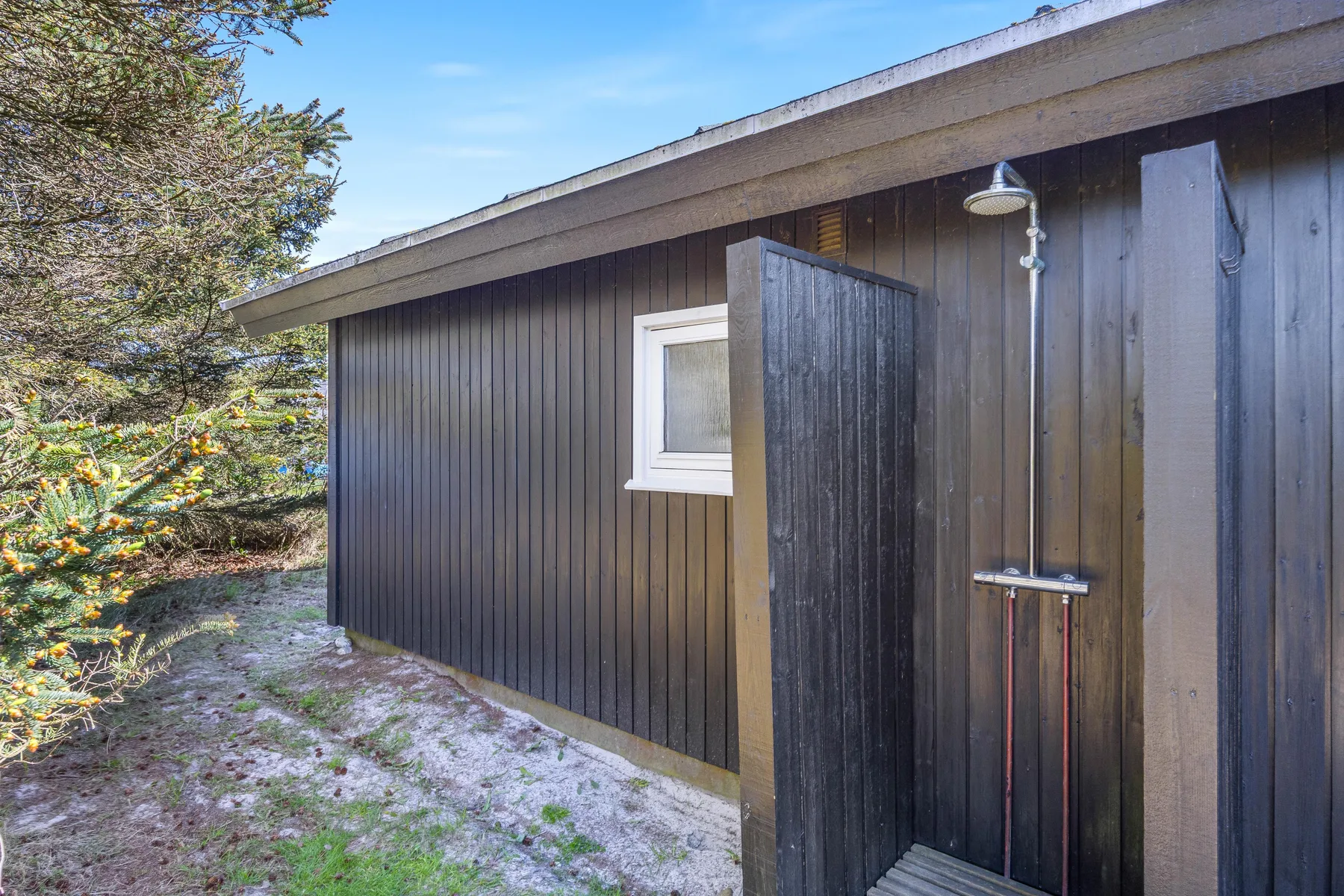 Exterior de una cabaña de madera oscura con una ventana pequeña y una ducha exterior.