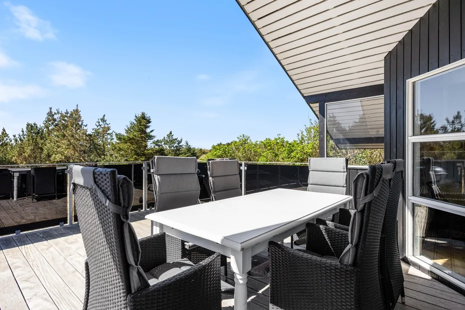 Una terraza exterior con mesa de comedor y asientos oscuros de tejido entrelazado da a unos árboles.