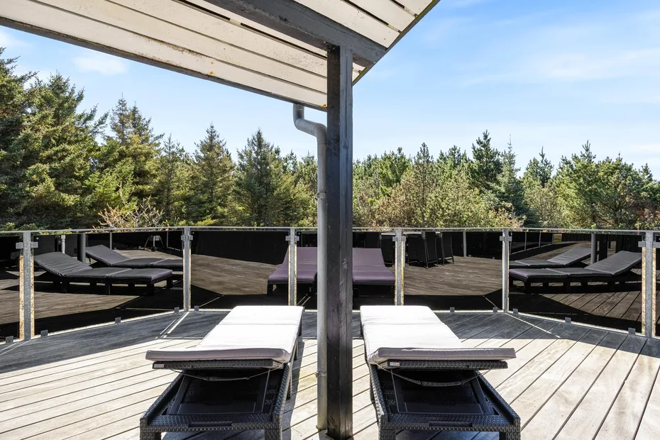Una terraza de madera con tumbonas da a una hilera de árboles perennes.