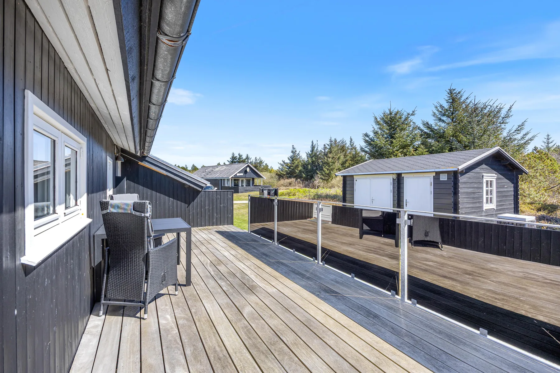 Una terraza de madera junto a una casa de revestimiento oscuro, con asientos al aire libre y un patio cercado.
