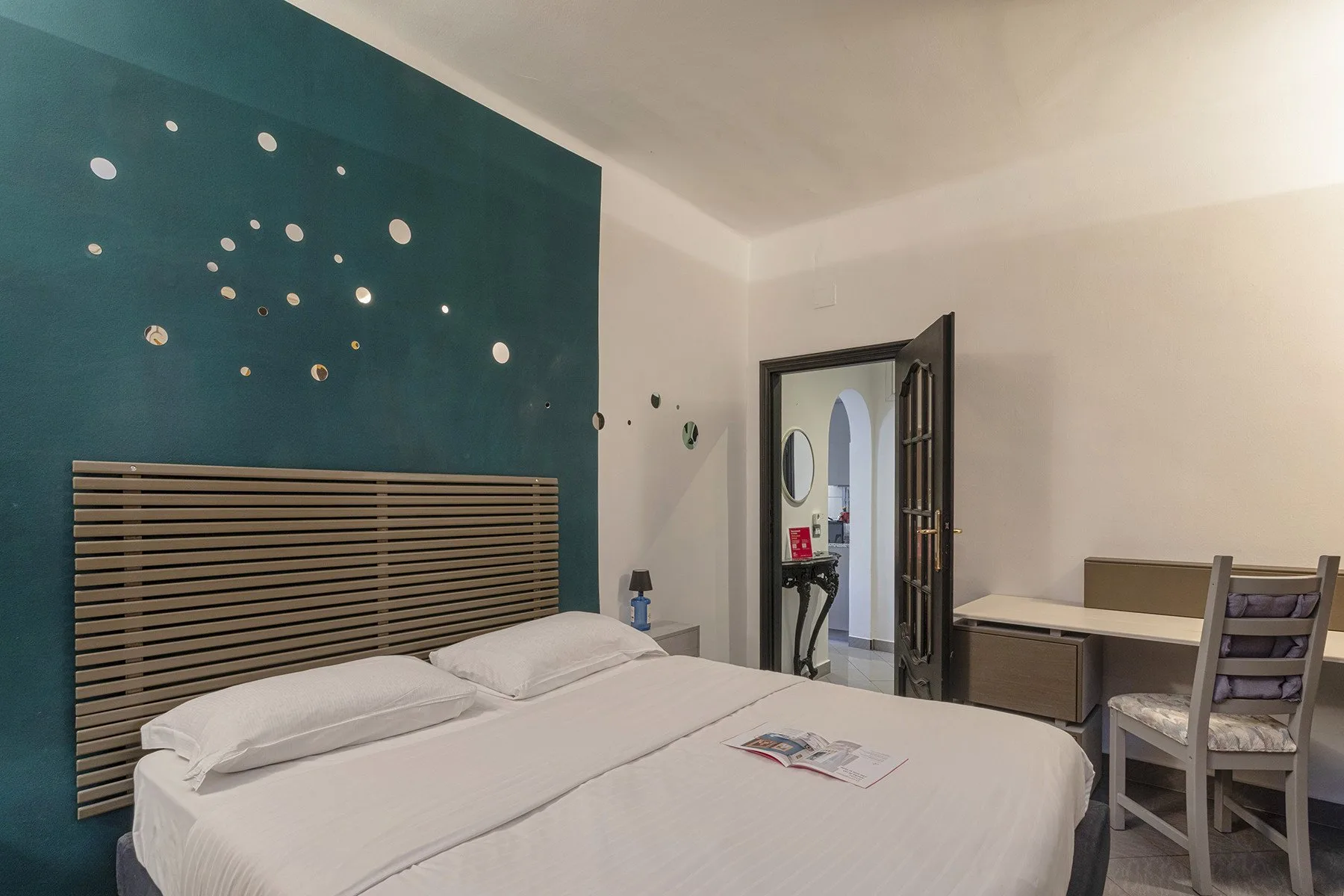 Un dormitorio con una cama junto a una pared decorativa verde azulado y un escritorio junto a la puerta.