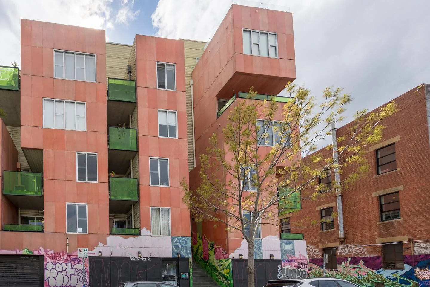 A modern apartment building with red facade panels and balconies stands beside a brick building.