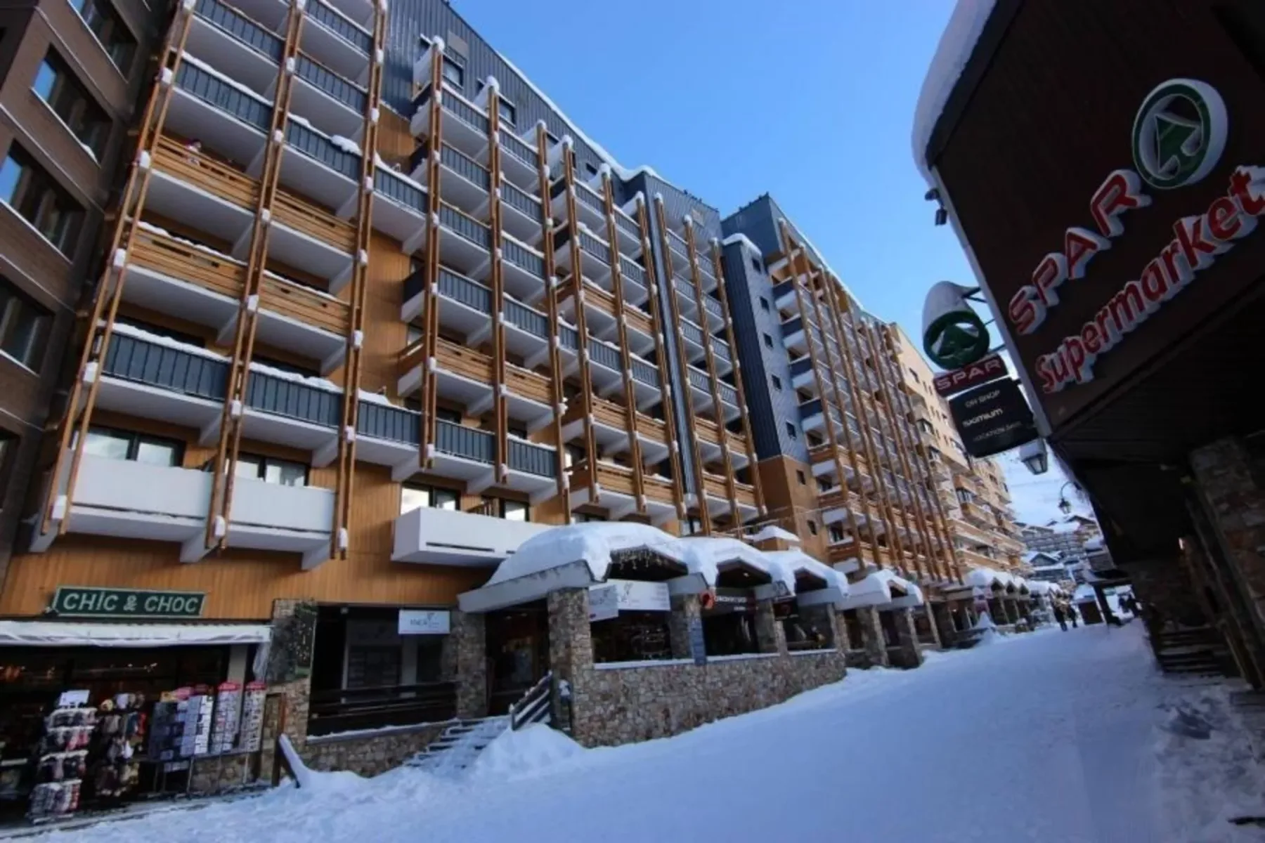 Calle cubierta de nieve junto a un edificio de varios pisos con balcones y locales.