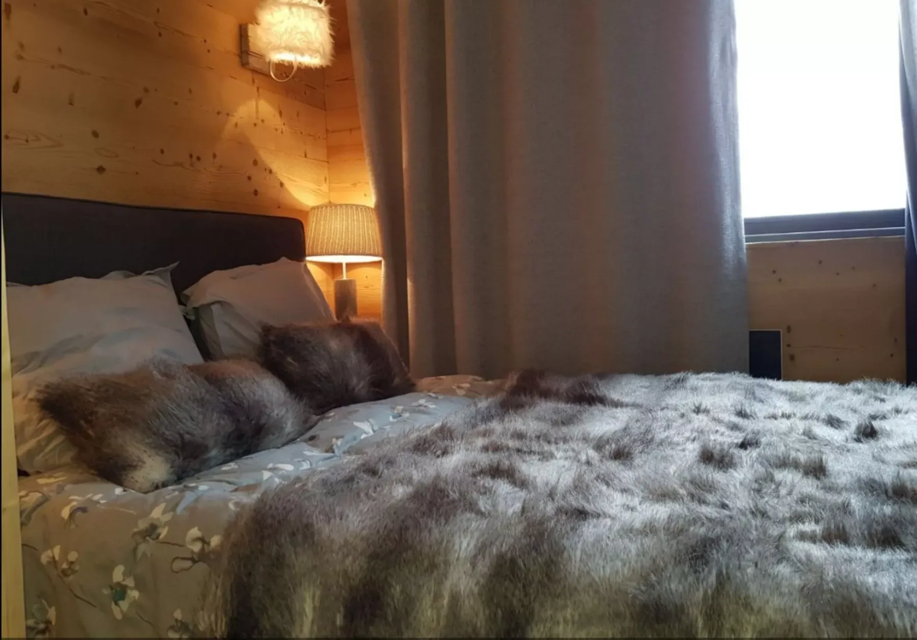 Un dormitorio con una cama cubierta por una manta gris texturizada junto a una ventana con cortinas.