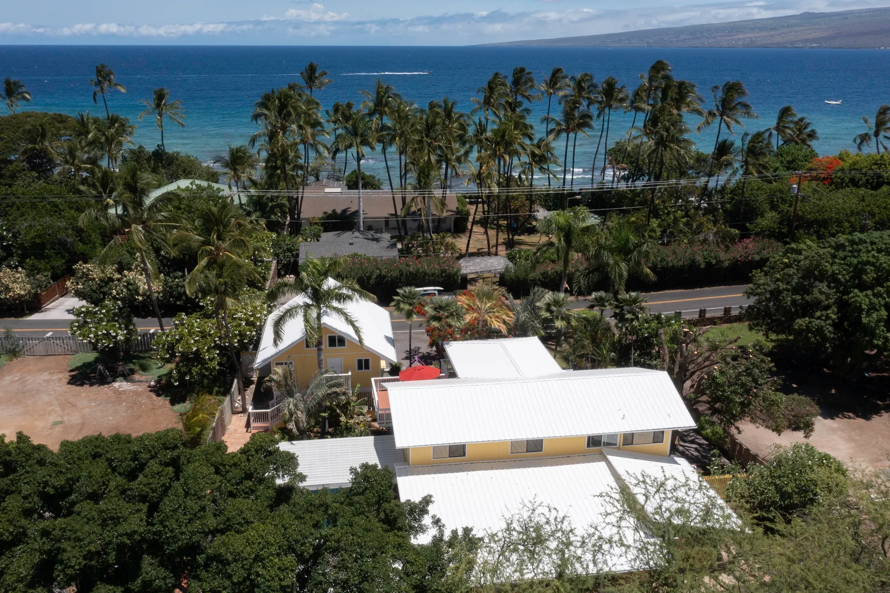 An aerial view of low buildings among tropical greenery, with palm trees and the ocean beyond.