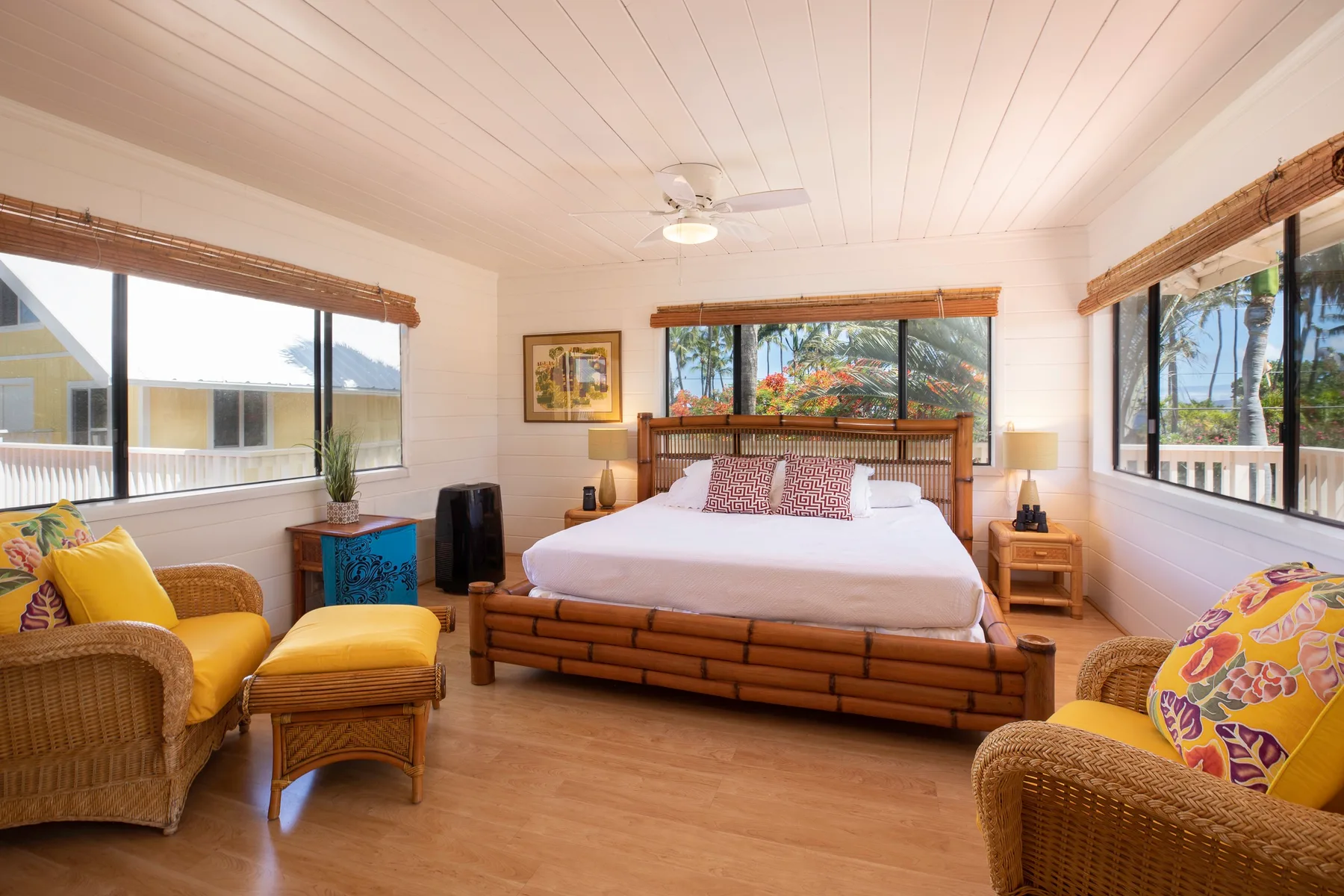 A bright bedroom with a large bed and wicker seating beside wide windows.