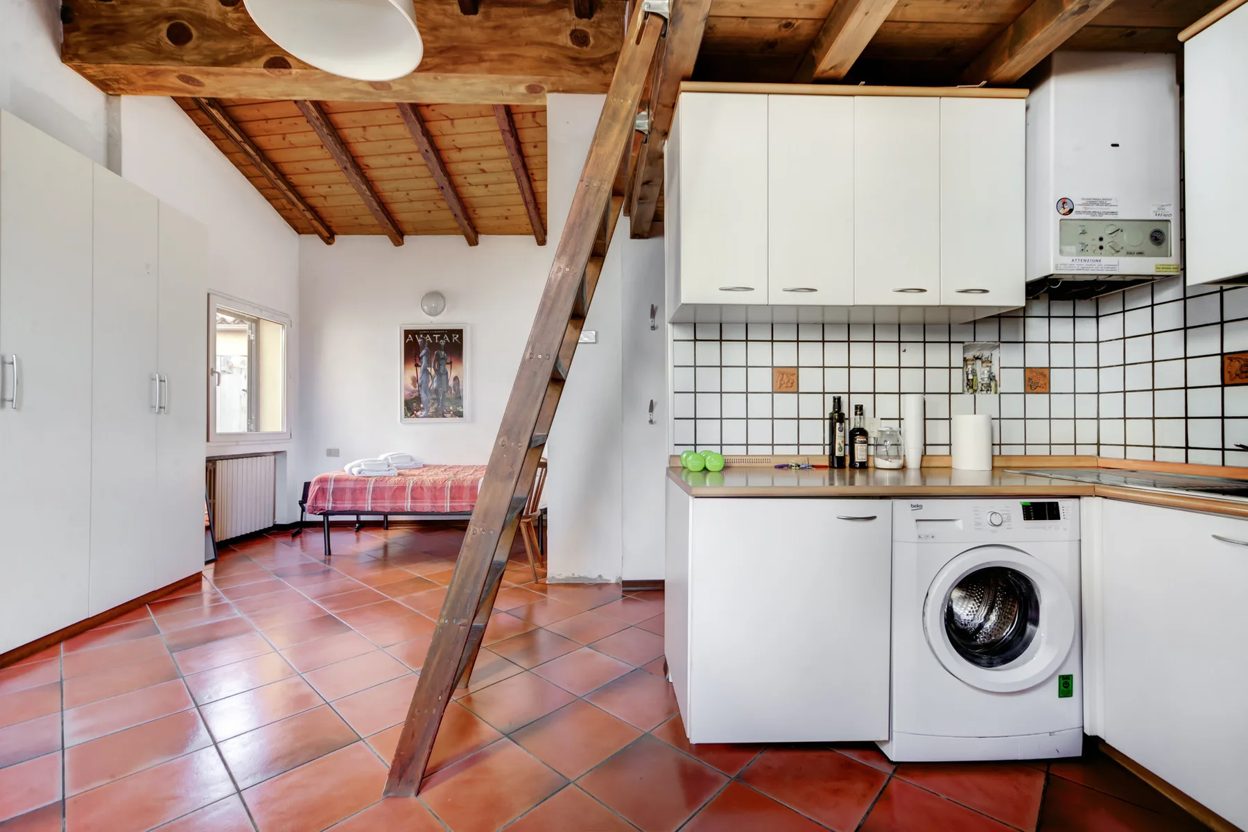 Una cocina pequeña con armarios blancos y una lavadora, junto a un dormitorio pequeño.