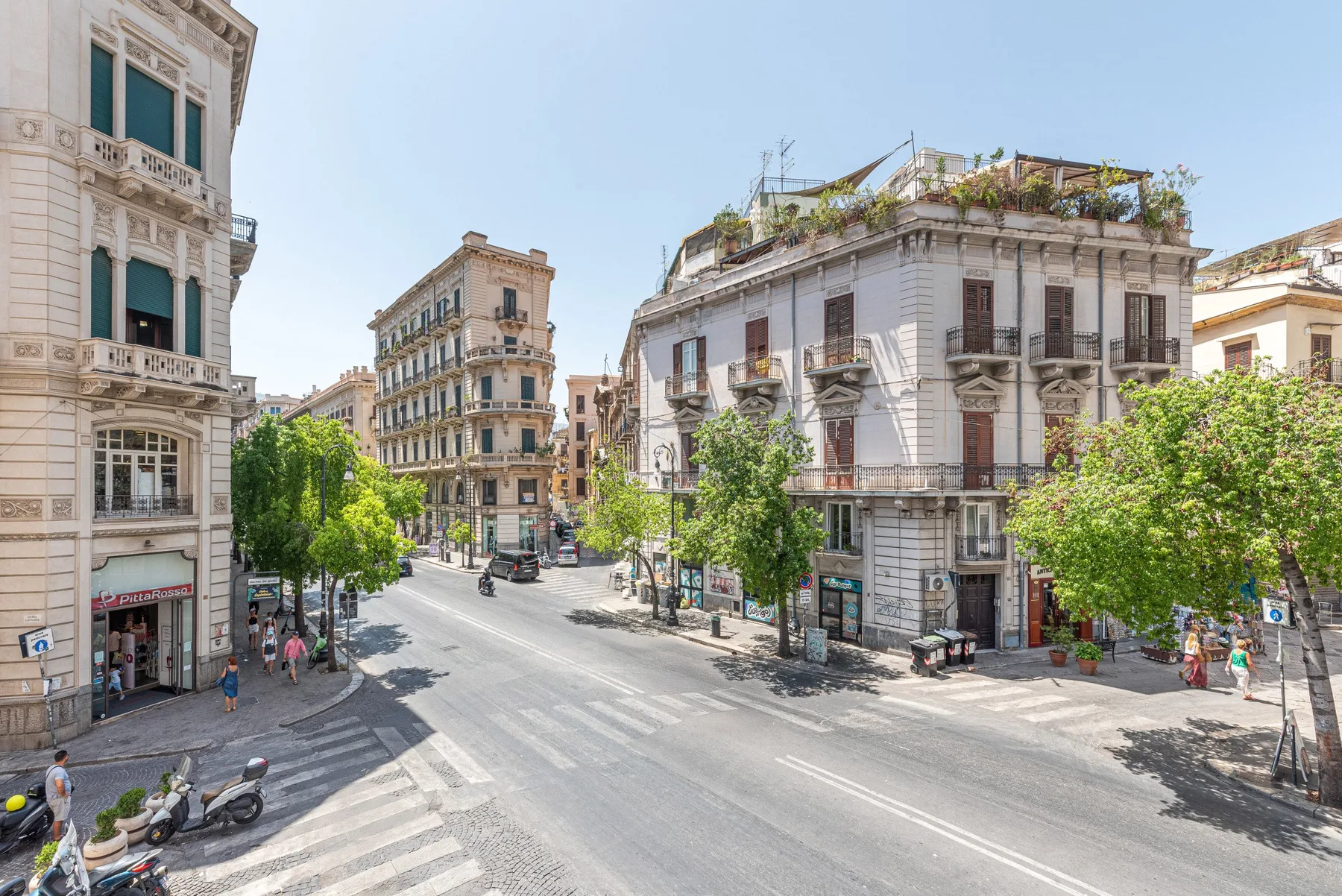 An urban intersection lined with ornate multi-story buildings and leafy street trees.