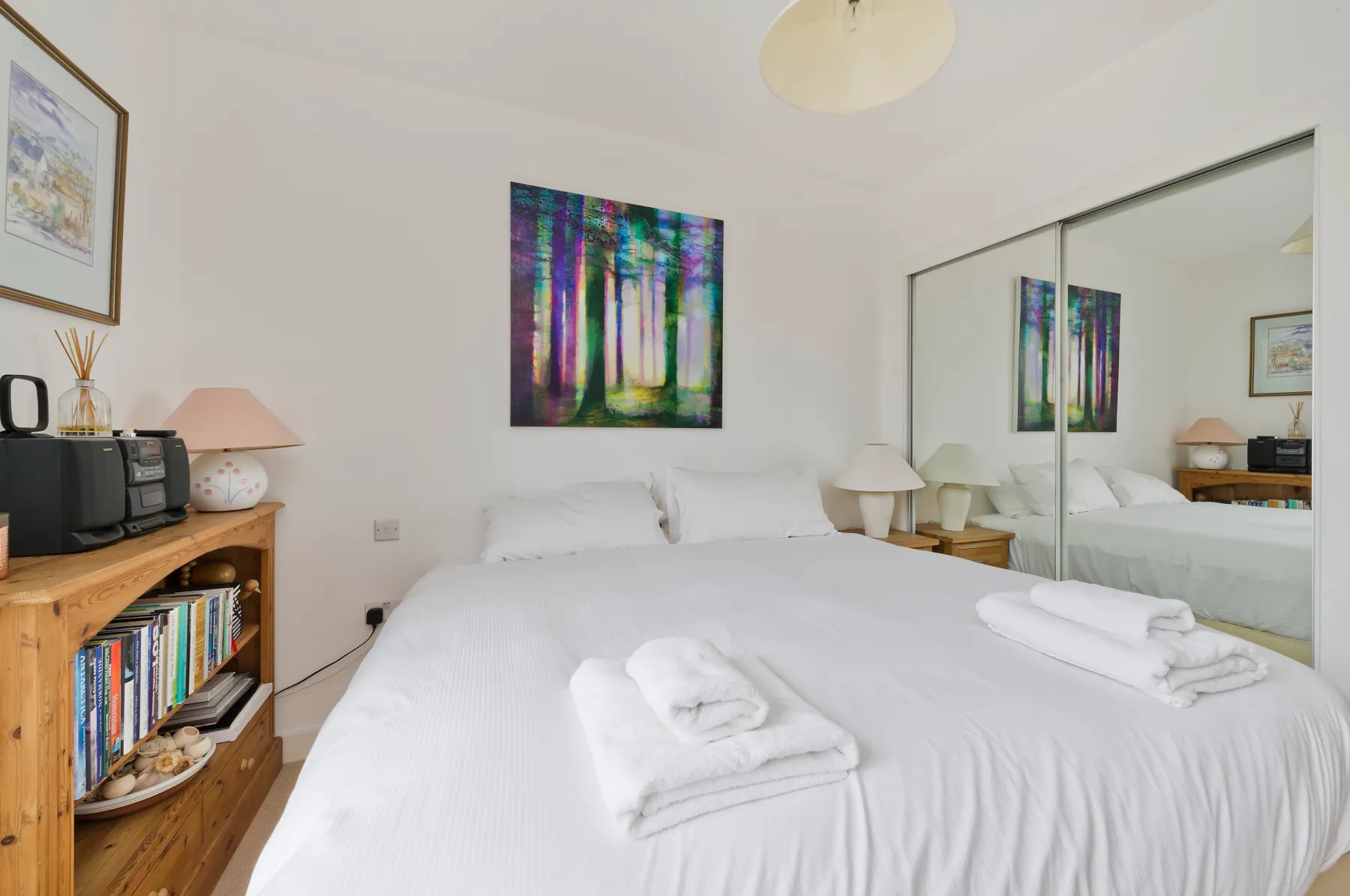 Un dormitorio luminoso con ropa de cama blanca y un cuadro colorido de un bosque sobre la cama.