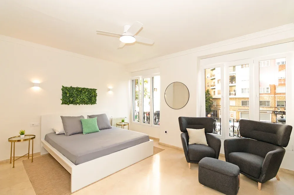A bright bedroom with a low bed and two dark armchairs beside large windows.