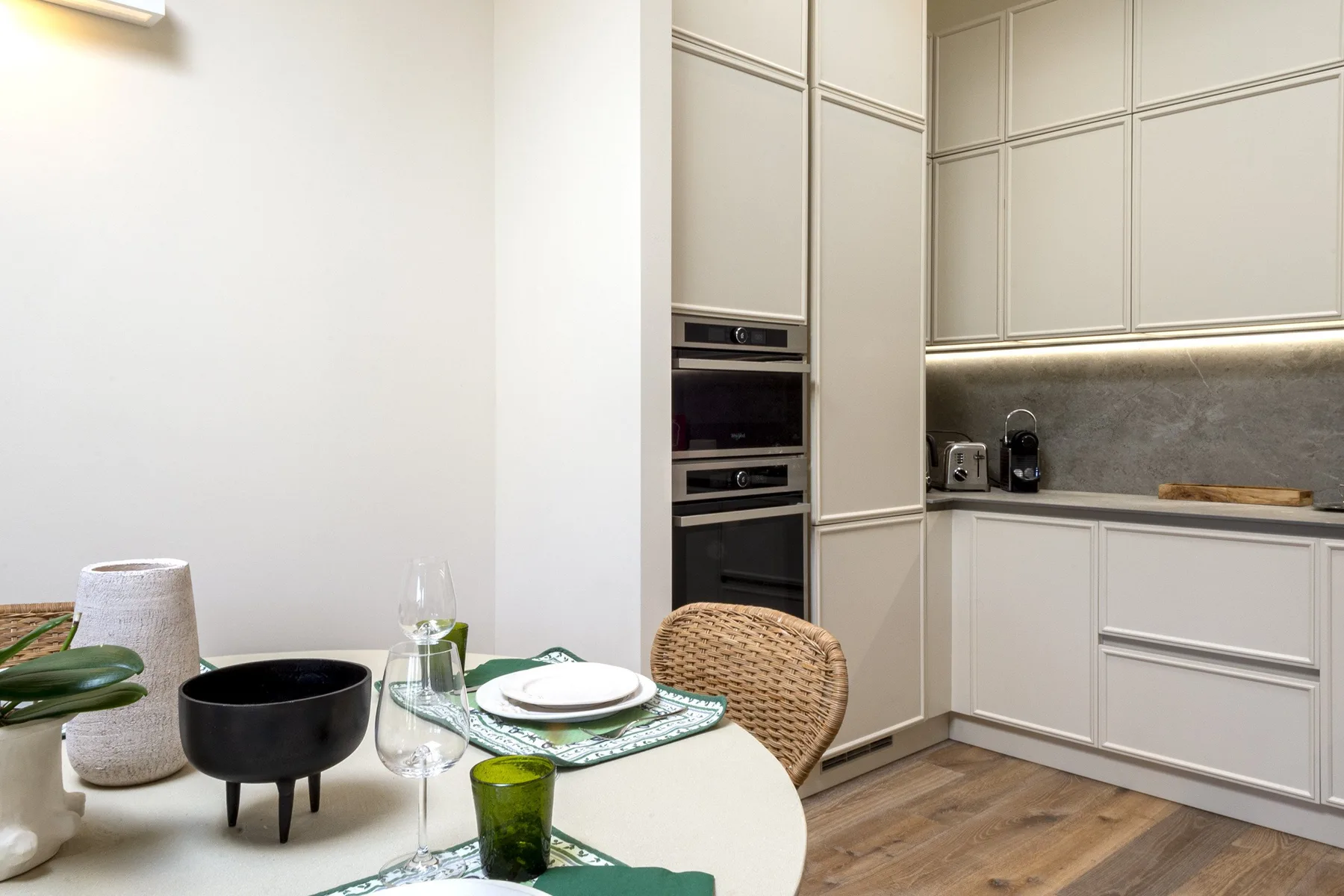 A dining area beside a white kitchen, with a set table and built-in oven.