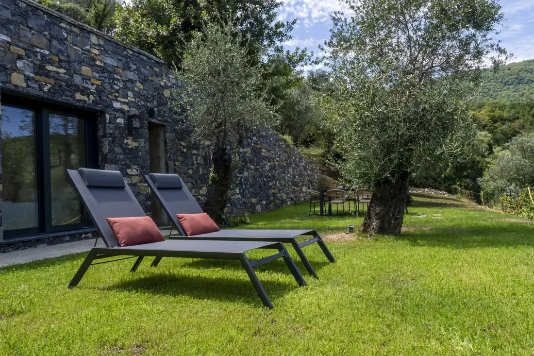 A grassy outdoor seating area beside a stone building, with trees and loungers.