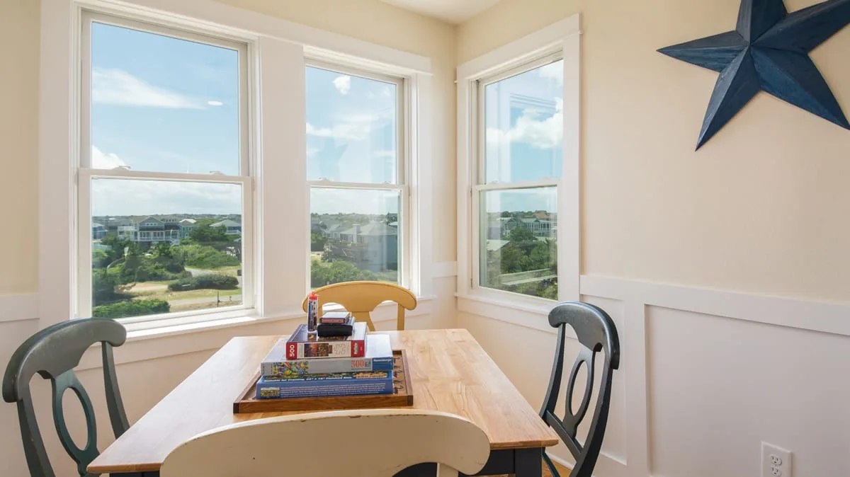 A small dining area with a wooden table beside three large windows.