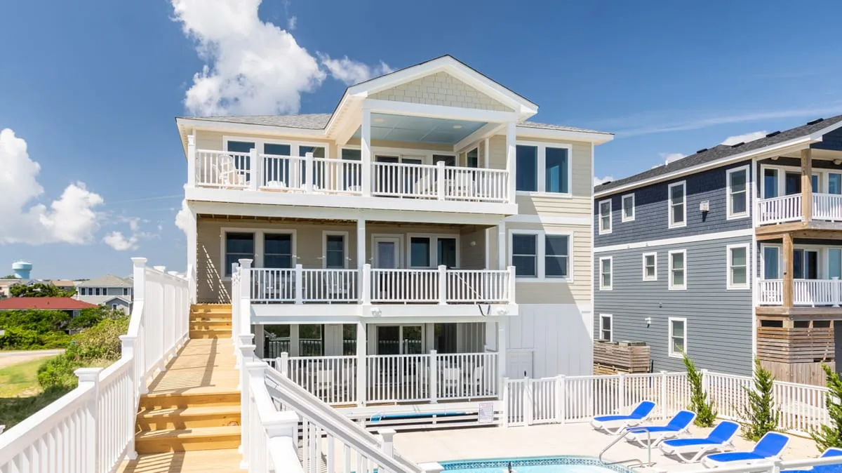 A large coastal-style house with balconies overlooks an outdoor pool and seating area.