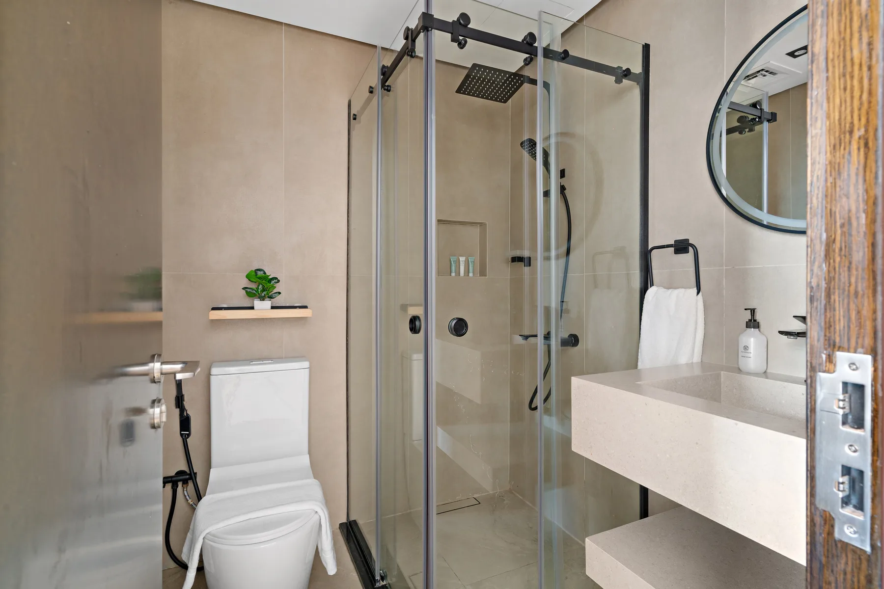 Un baño con ducha de vidrio, inodoro y lavabo bajo un espejo redondo.