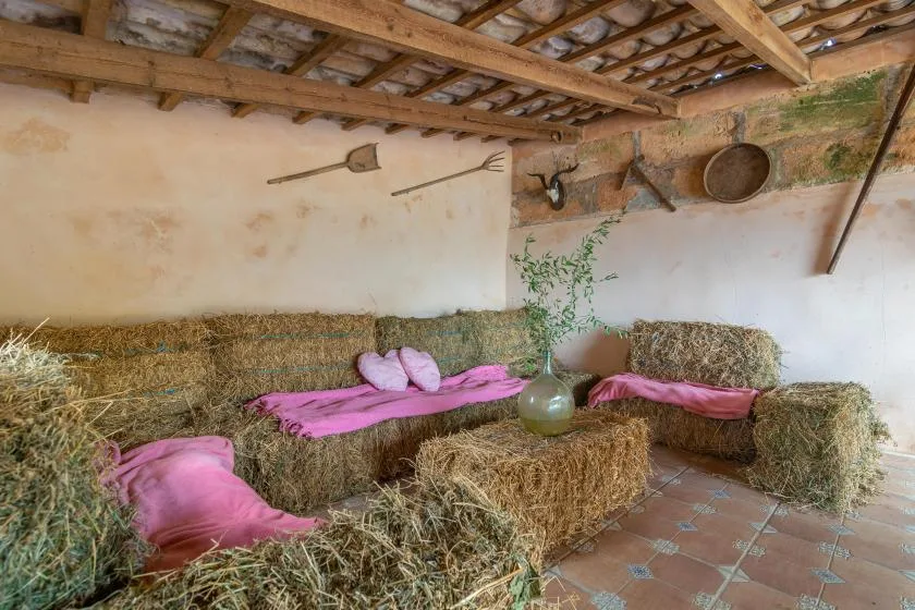 A rustic indoor seating area with hay bales and pink cushions beneath exposed wooden beams.