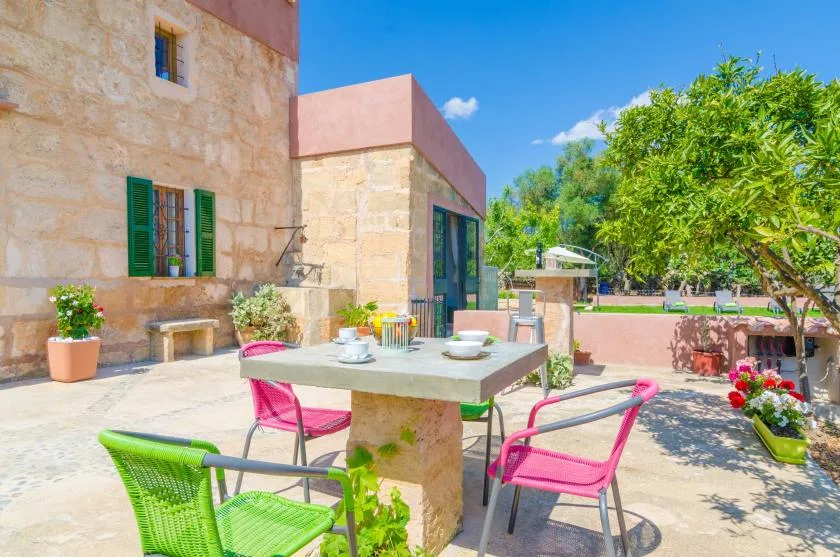 A sunny outdoor dining patio with a stone table, colorful chairs, and surrounding trees.