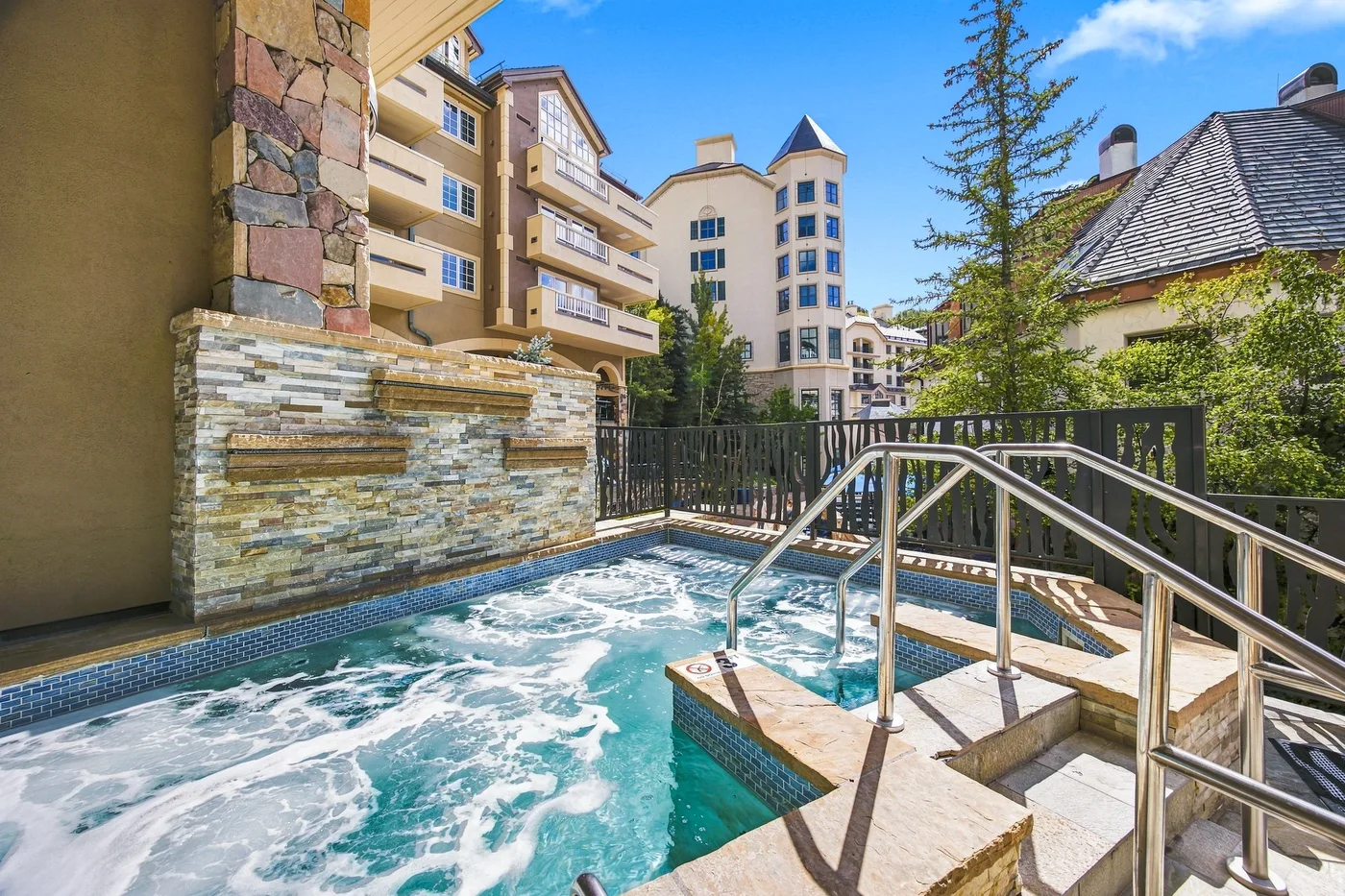 Una piscina exterior con agua burbujeante está junto a edificios de apartamentos y una barandilla metálica.