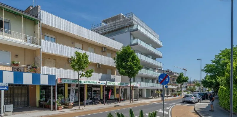A modern multi-story building stands along a sunny street with storefronts at ground level.