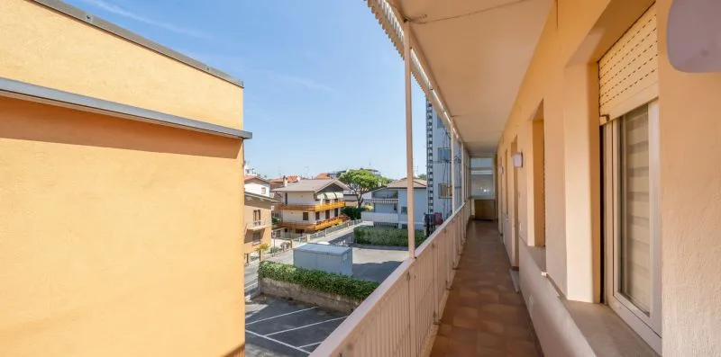 A long apartment balcony corridor overlooks nearby buildings and a parking area.