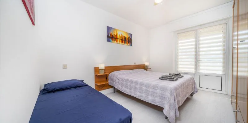 A bright bedroom with two beds and wide windows with blinds.