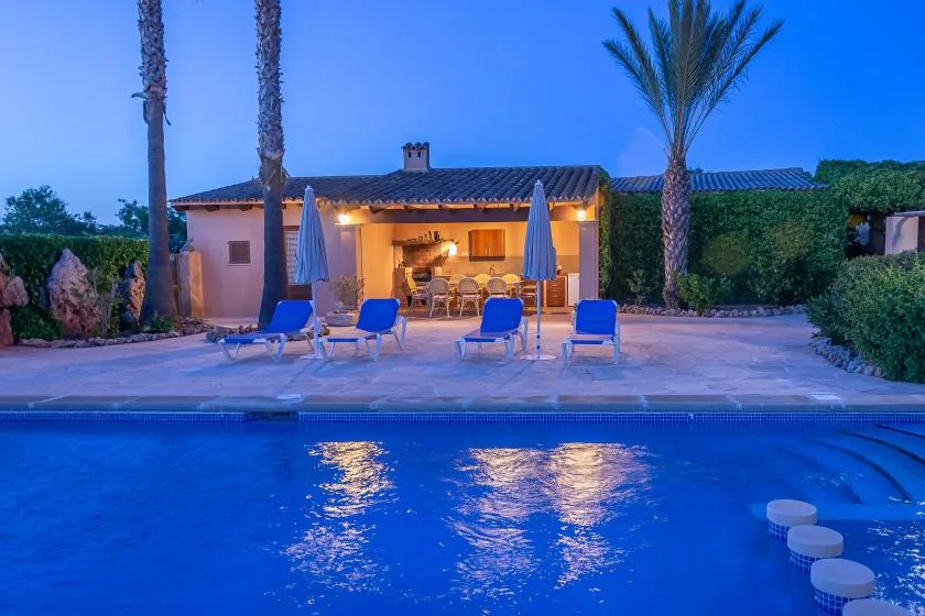 Una piscina da a un patio iluminado y una casa de una planta al anochecer.