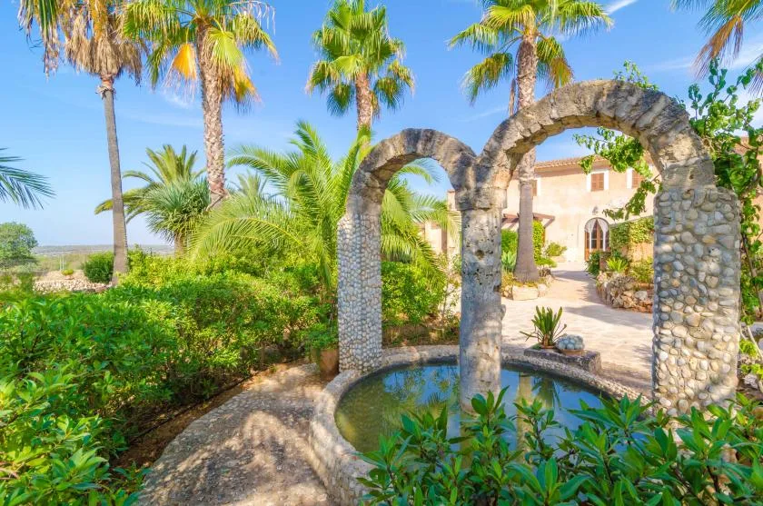 Un jardín paisajístico con arcos de piedra, palmeras y una pequeña piscina.