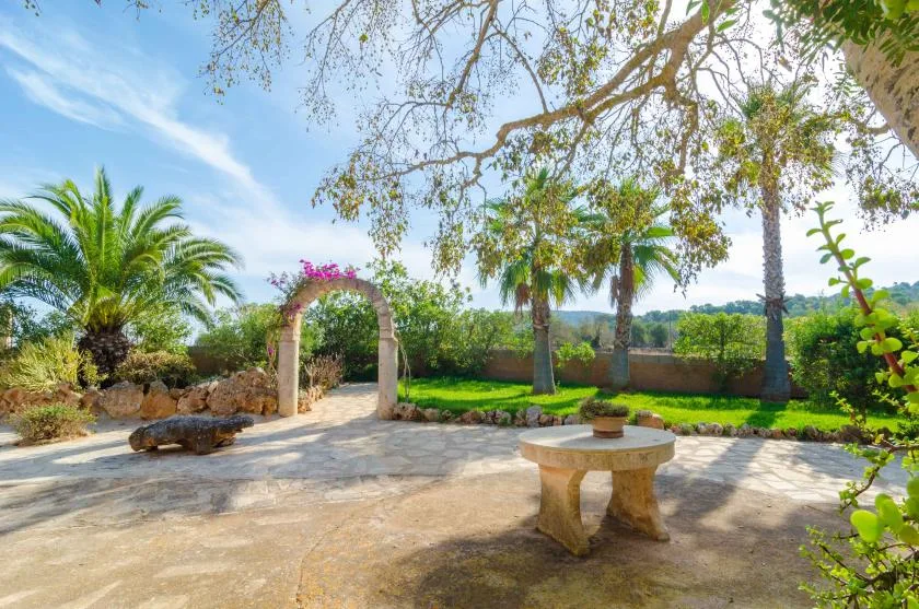 Un patio soleado con una mesa de piedra, palmeras y un arco con flores.