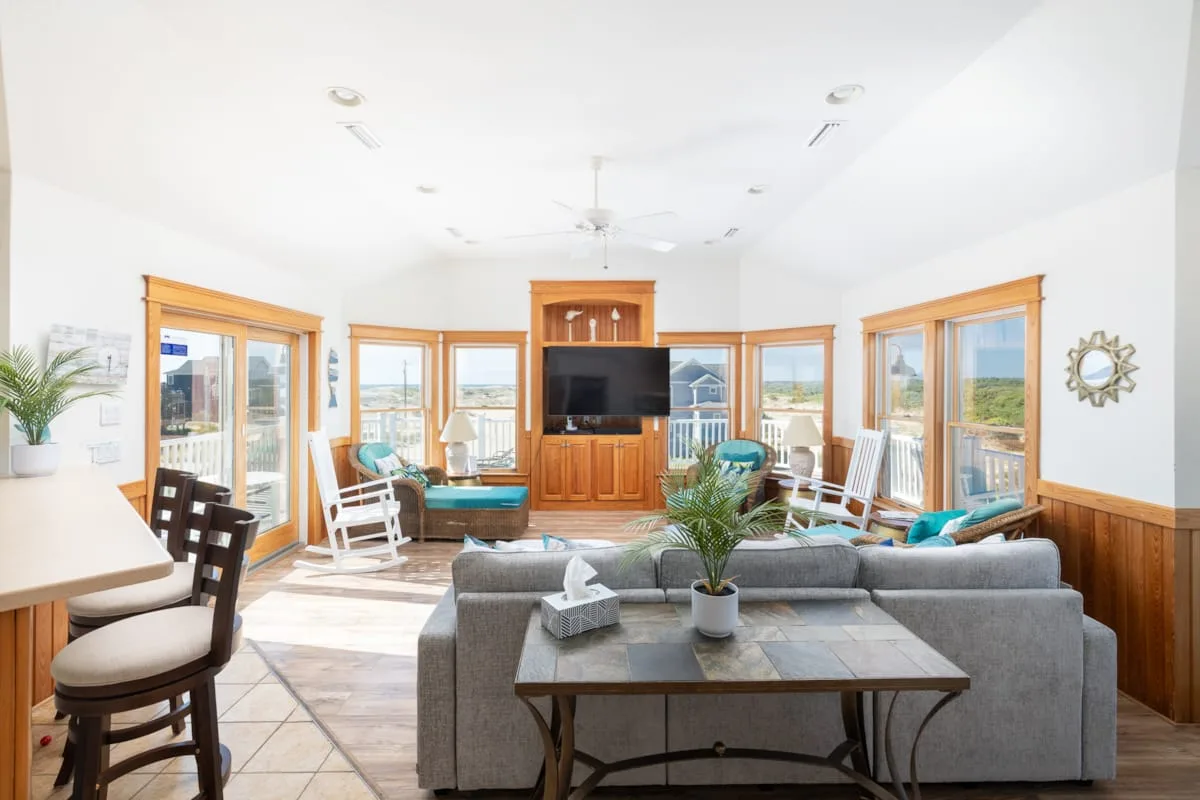 A bright living room with gray seating, a television, and large windows.