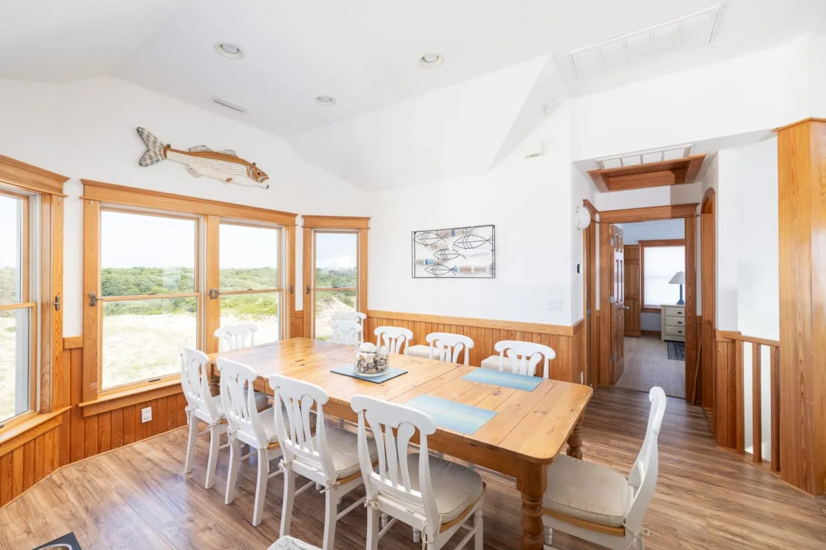 A bright dining room with a long wooden table beside large windows.