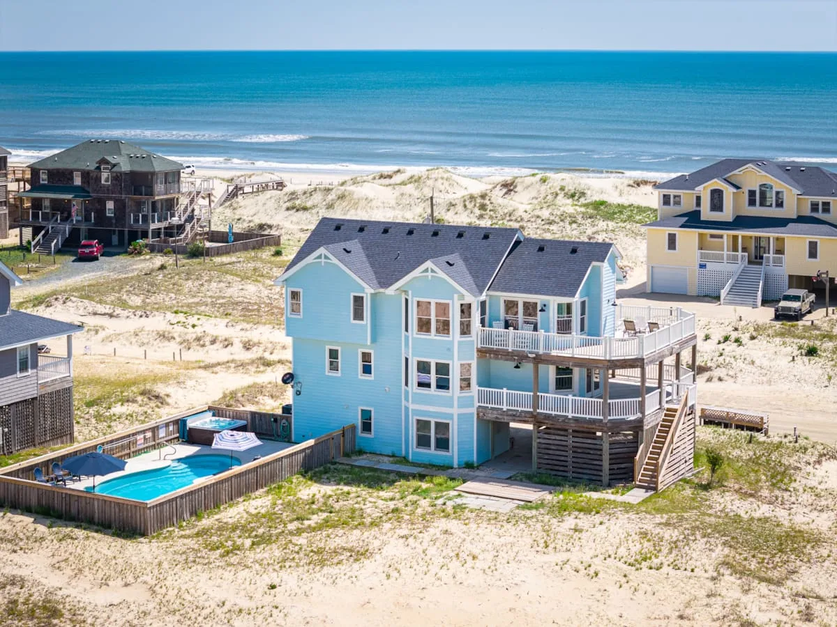 A blue beach house stands among sandy dunes beside the ocean.