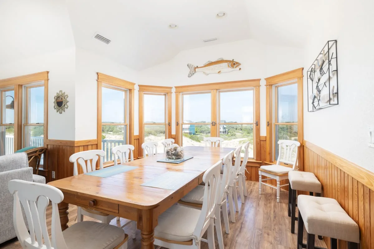 A bright dining room has a long wooden table surrounded by white seating.