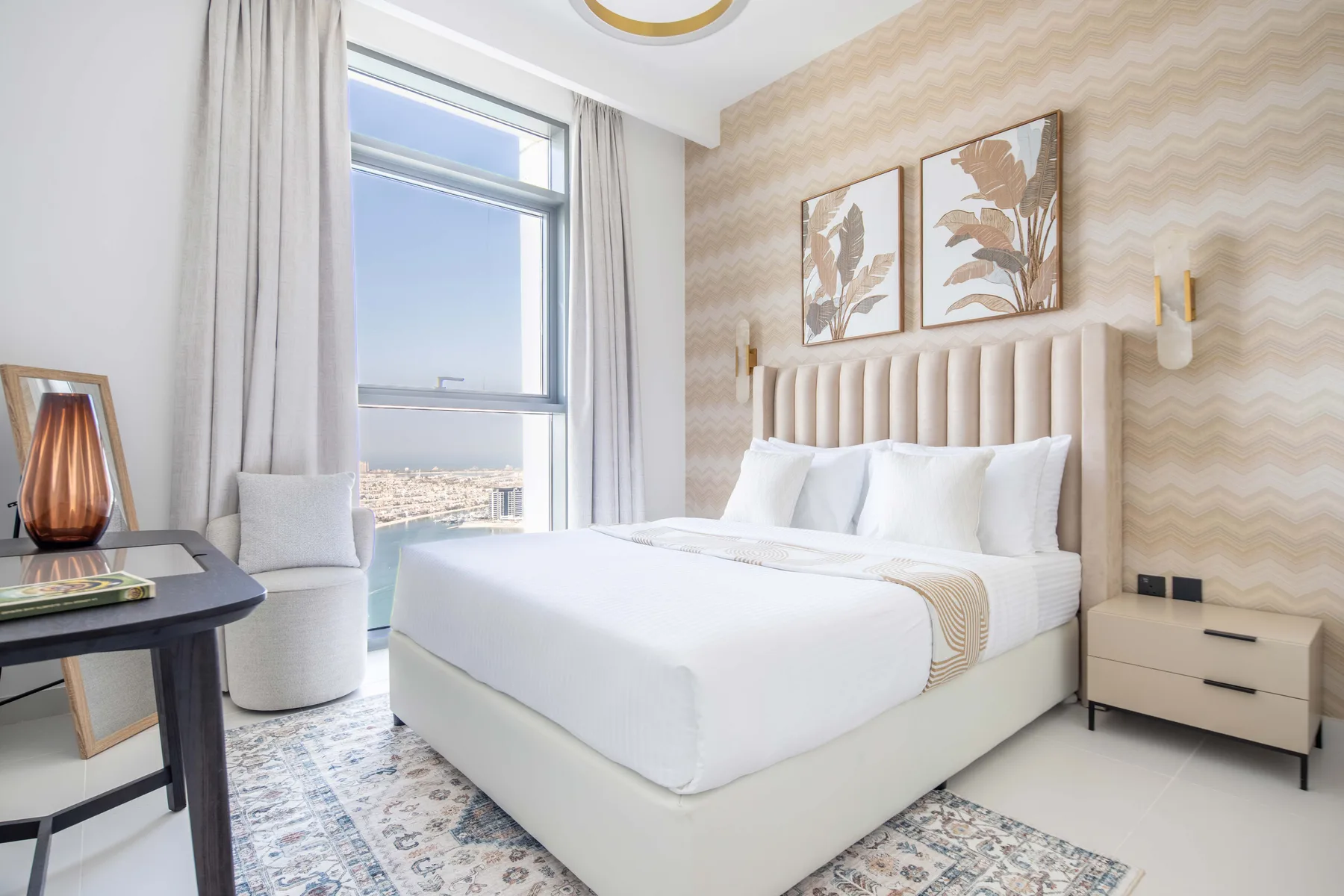 Un dormitorio luminoso con una cama bien tendida, papel tapiz estampado y una ventana grande.