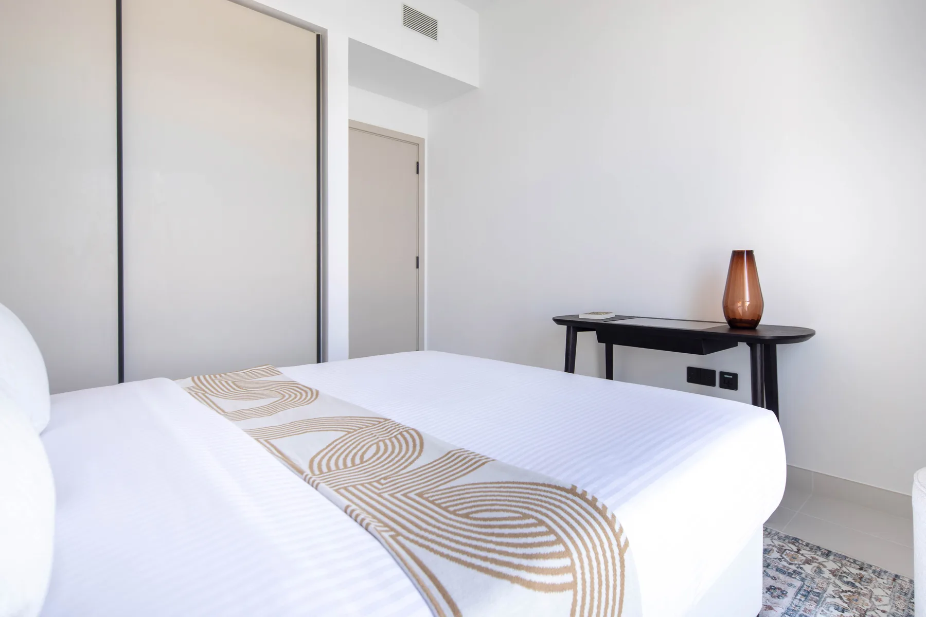 Dormitorio luminoso con una cama tendida y una mesa pequeña junto a la pared.