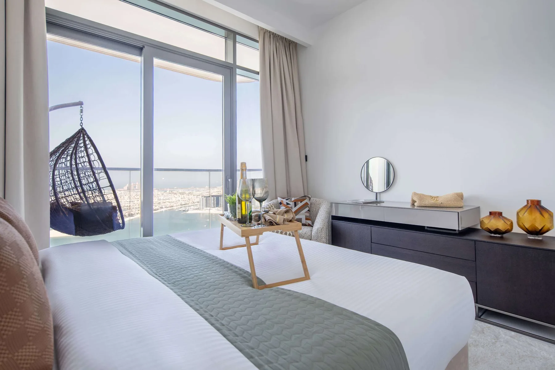 Un dormitorio luminoso con una cama tendida junto a grandes ventanales y una silla colgante.
