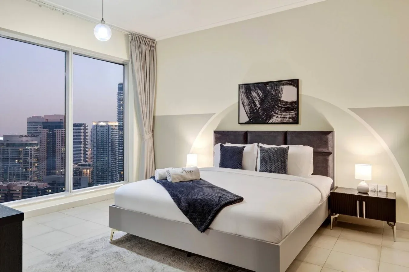 Un dormitorio con una cama tendida junto a grandes ventanales con vistas a edificios de la ciudad.
