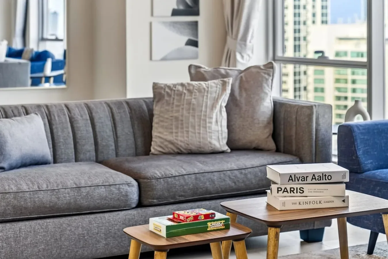 Una sala de estar con un sofá gris, cojines y mesas junto a una ventana.