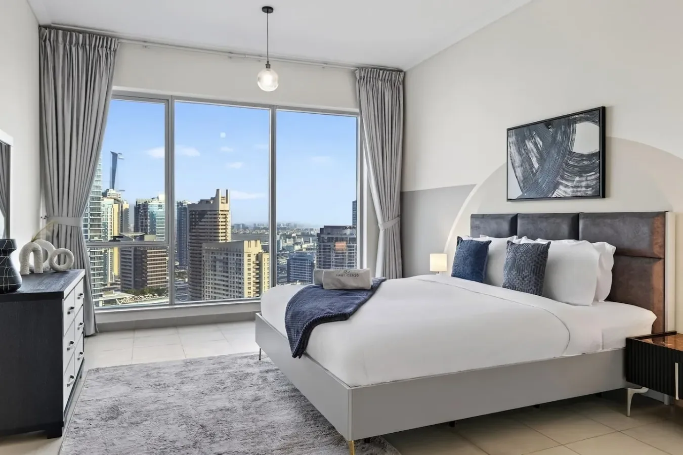 Un dormitorio luminoso con una cama bien tendida y grandes ventanas con vistas a edificios urbanos.