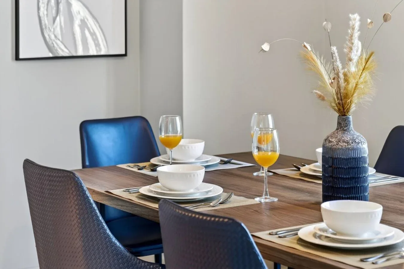 Una mesa de comedor está puesta con platos, copas y un jarrón con flores secas.