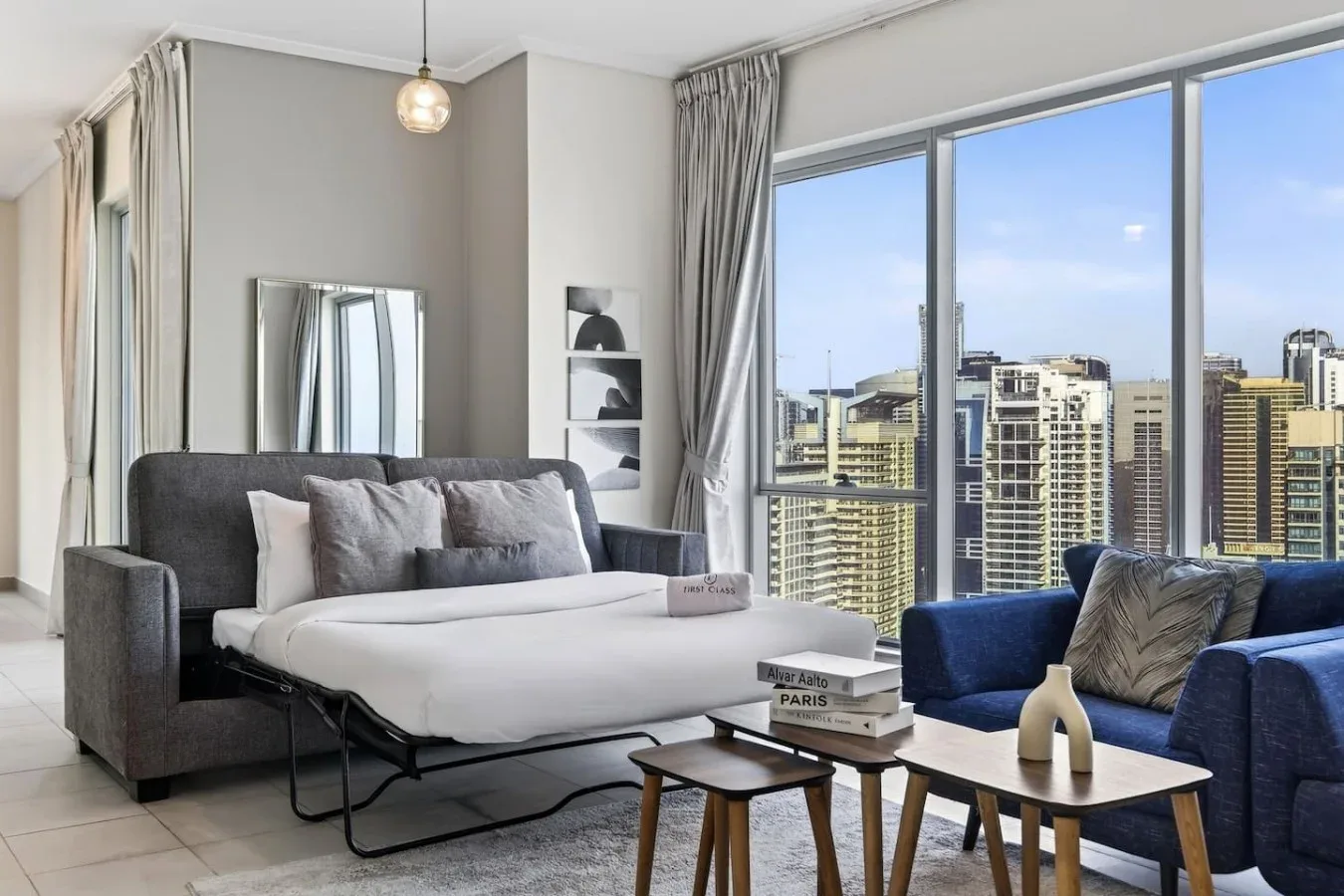 Un dormitorio con una cama ordenada y asientos junto a grandes ventanales con vistas a la ciudad.