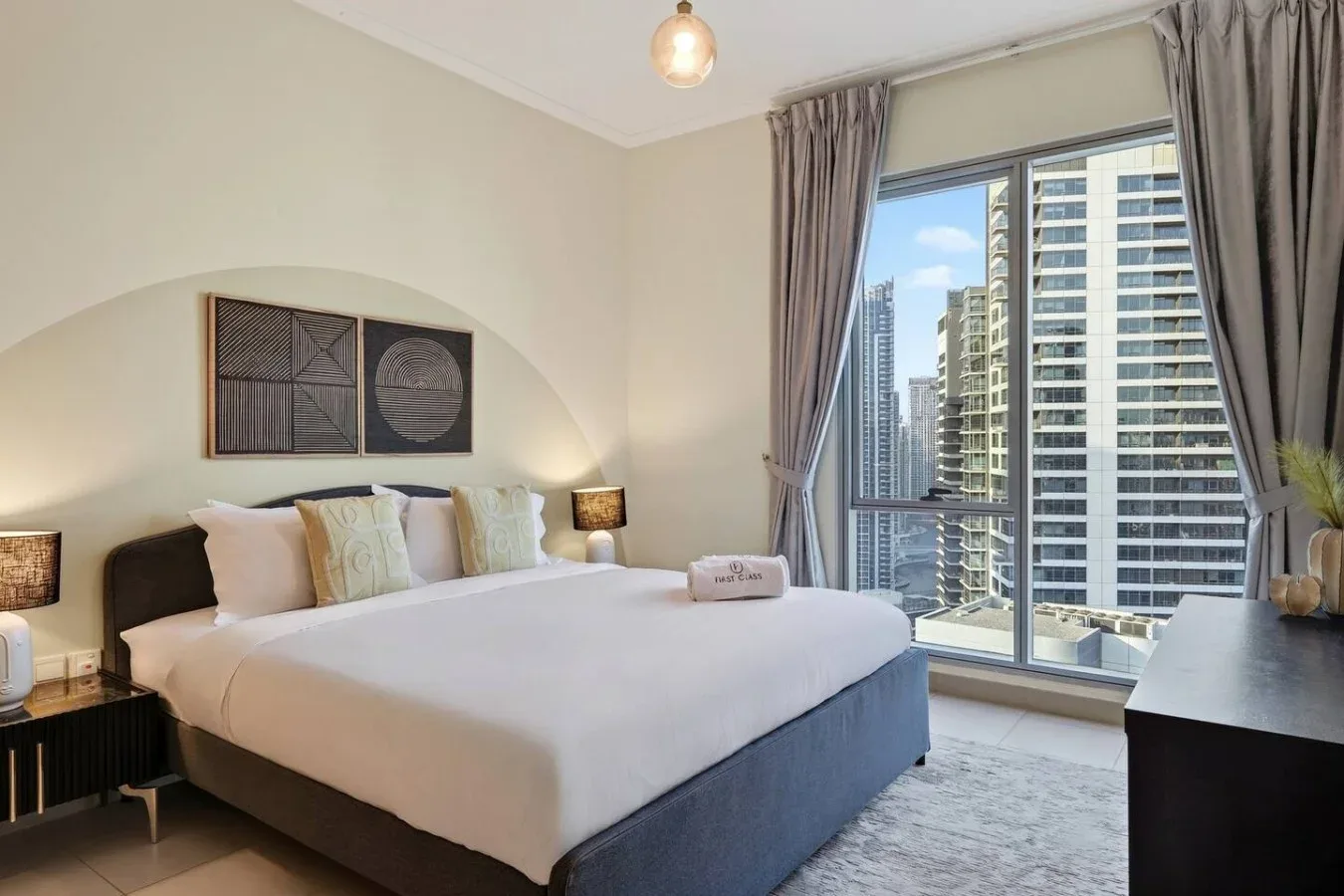 Un dormitorio con una cama bien arreglada junto a una gran ventana con edificios a la vista.
