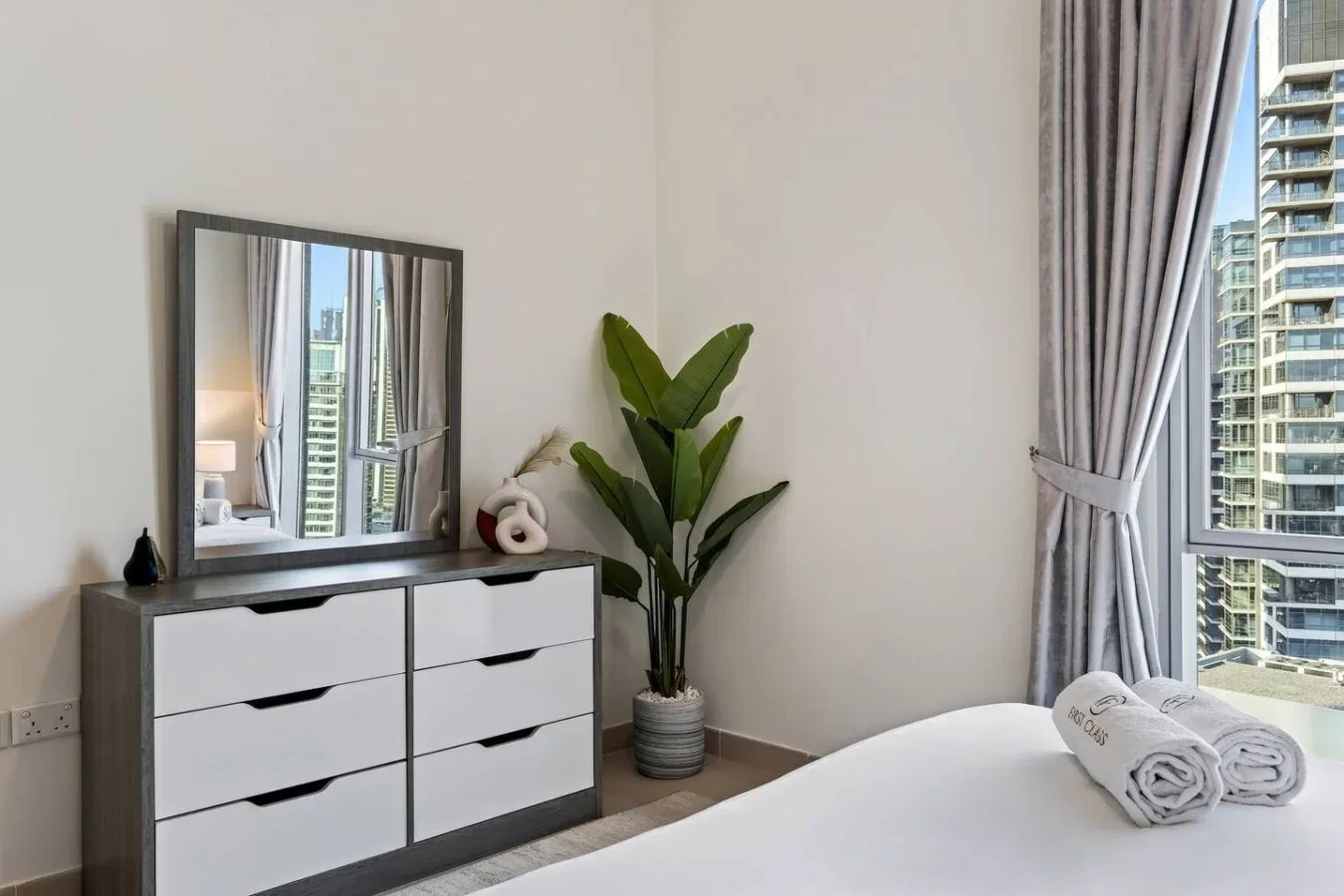 Un dormitorio luminoso con una cómoda, una planta frondosa y una ventana amplia con edificios a la vista.